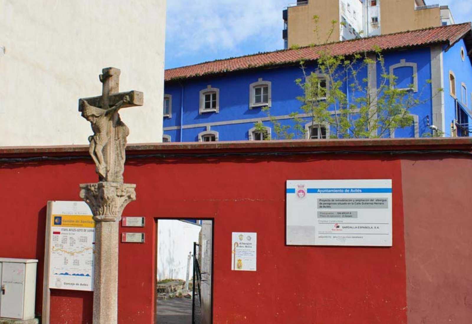 Entrée de l'auberge de pèlerins avec une croix de pierre et un mur rouge près d'un bâtiment bleu sur l'étape de Gijón à Avilés du Chemin du Nord.