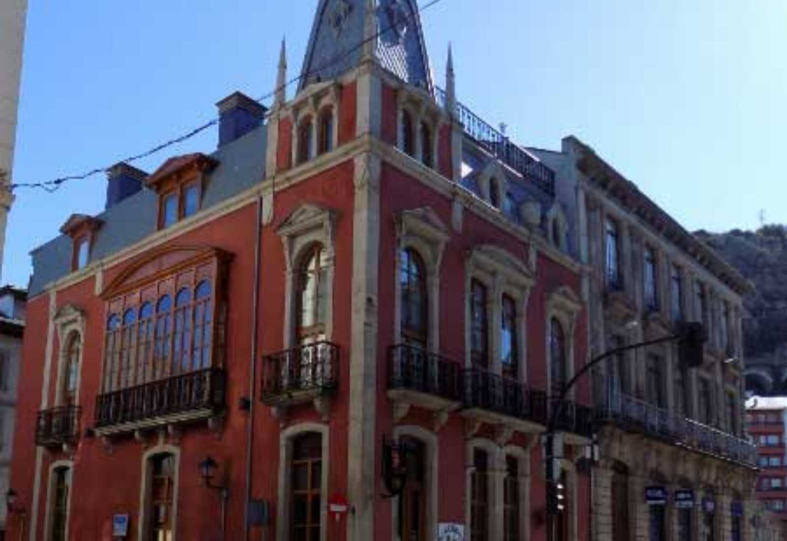 Casarão modernista avermelhado com pináculo de ardósia e galerias envidraçadas numa esquina na etapa de Luarca a La Caridad do Caminho do Norte.