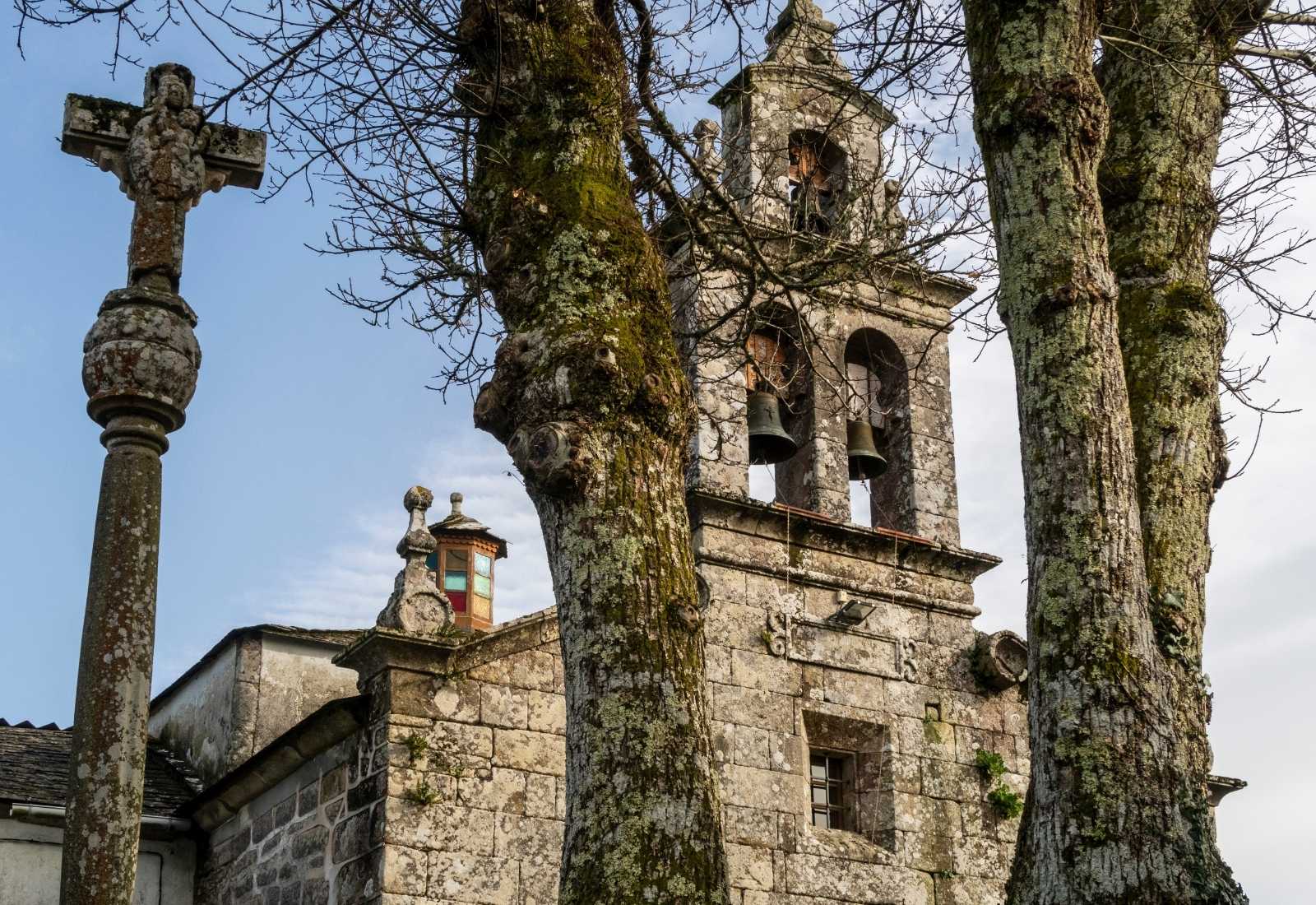 Église de pierre de Miraz au clocher-mur à deux cloches et un cruceiro entre des arbres nus, étape De Baamonde à Miraz du Chemin du Nord.
