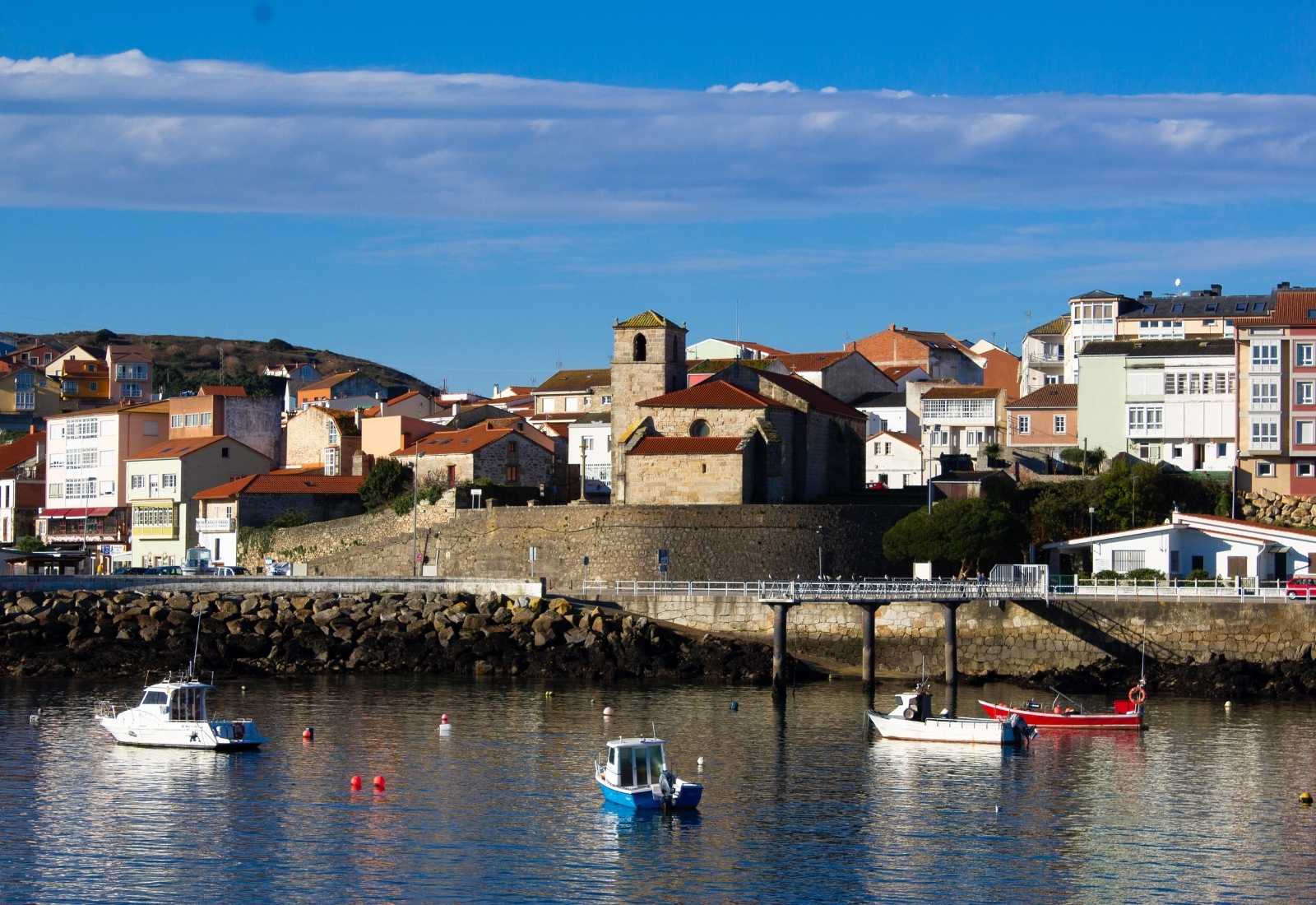 Puerto de Camariñas con barcas fondeadas y la iglesia de piedra entre las casas bajo un cielo azul, etapa de Camariñas a Muxía.