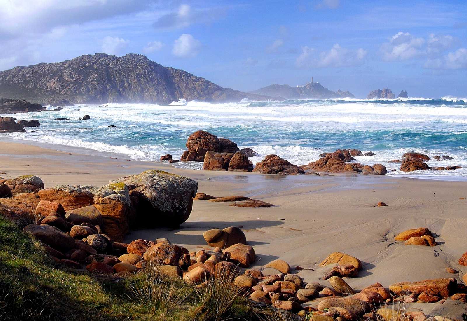 Playa salvaje con rocas anaranjadas y olas rompiendo bajo un cabo lejano en la etapa de Arou a Camariñas del Camino dos Faros