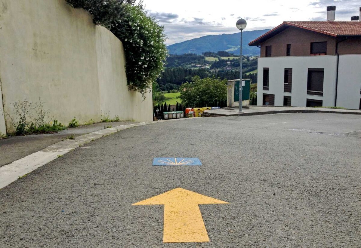 Freccia gialla e conchiglia dipinte sull'asfalto che segnano il percorso, col paese sullo sfondo, tappa Da Deba a Markina del Cammino del Nord.