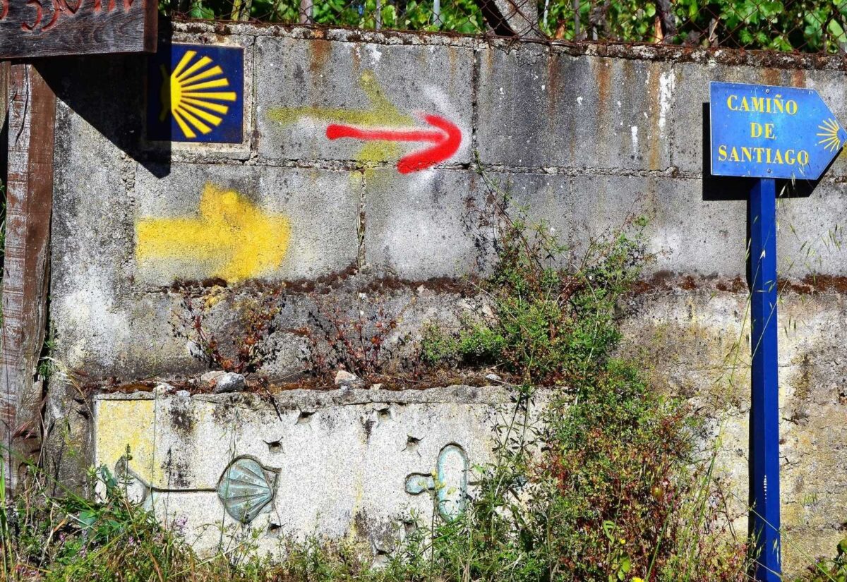 Wegmarkierung des Camino mit Muschel, gelben Pfeilen und blauem Schild an einer Mauer auf der Etappe Von A Gudiña nach Laza.