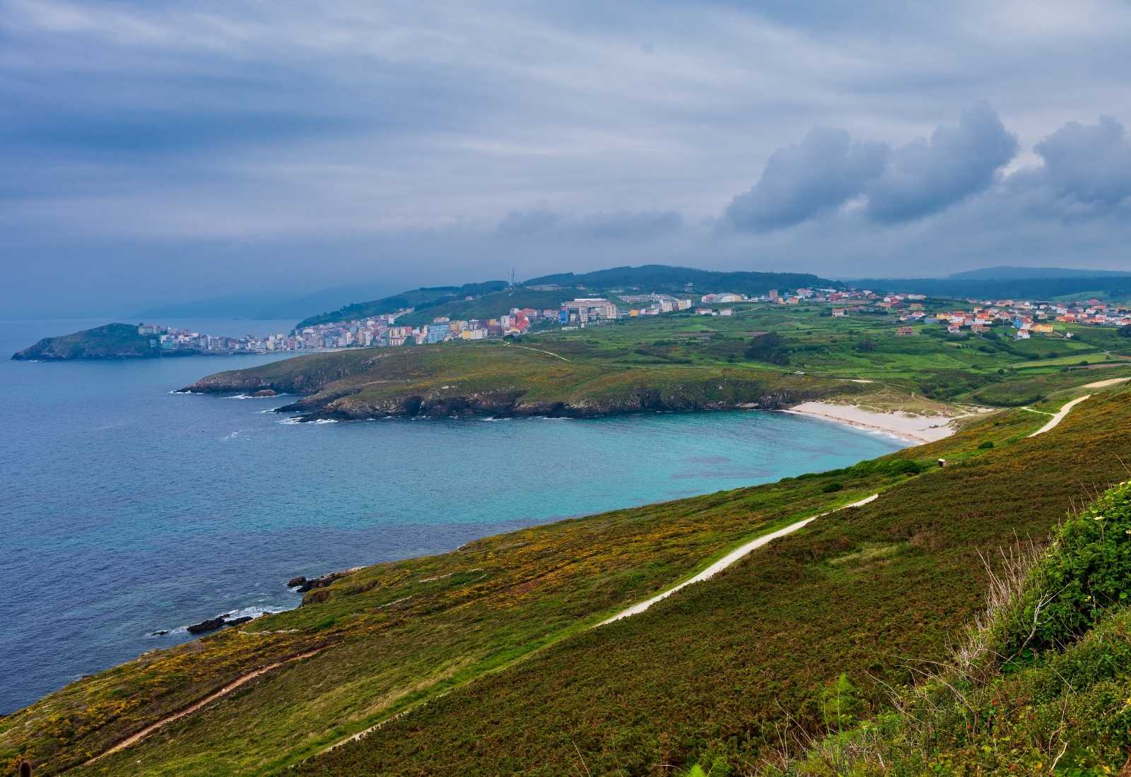 Vista elevada del pueblo y su cala turquesa entre laderas de brezo a la llegada a Malpica de Bergantiños en el Camino dos Faros