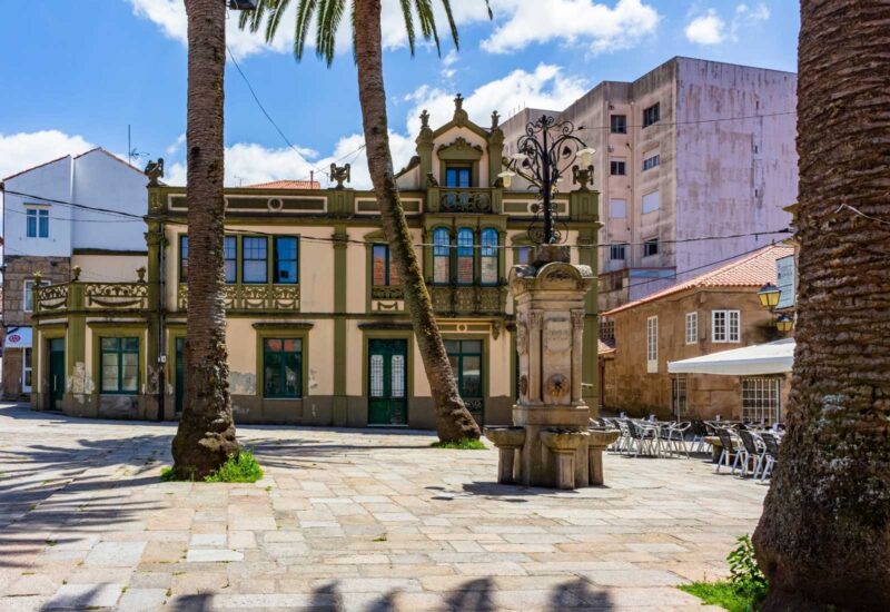 Praça com palmeiras e uma fonte de pedra diante de uma fachada modernista na etapa de O Grove a Cambados da rota do Padre Sarmiento