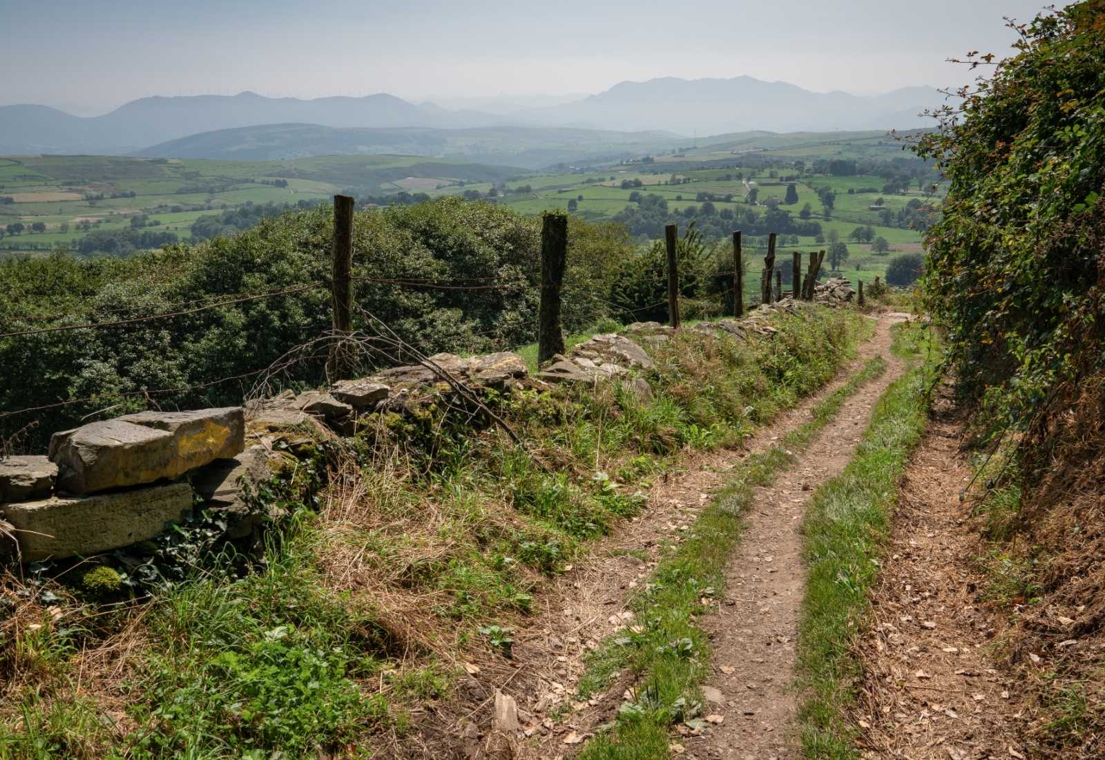 Trilho junto a um muro de pedra com vista sobre vales verdes, etapa de Pola de Allande a La Mesa do Caminho Primitivo