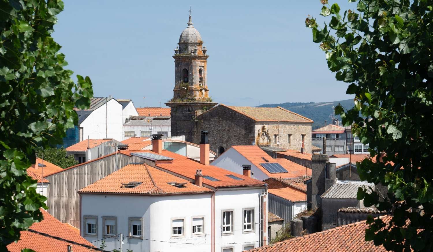 Telhados vermelhos de uma vila com a torre barroca de uma igreja entre a folhagem na etapa de Ferreira a Melide do Caminho Primitivo
