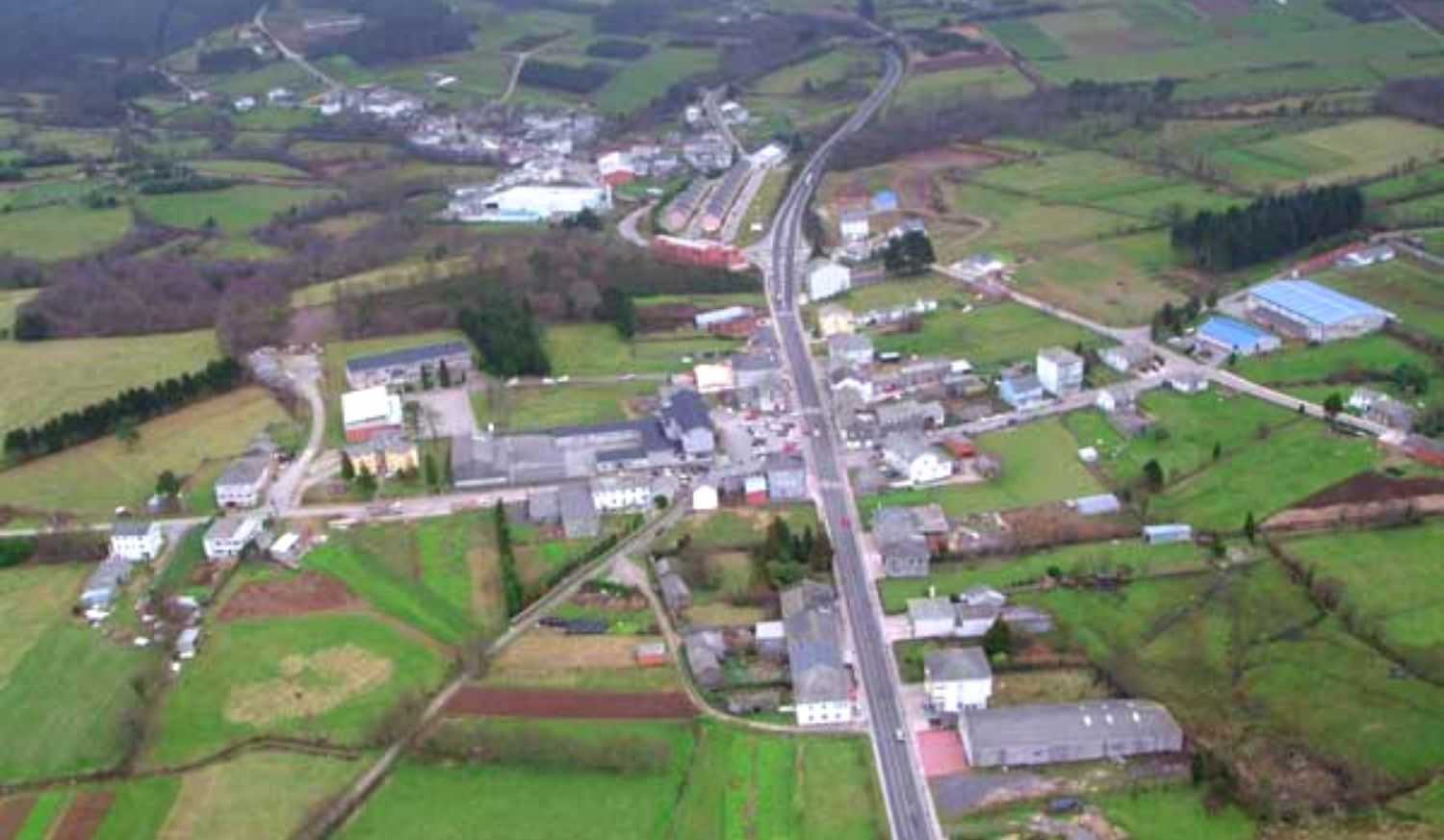 Vue aérienne du village d'Abadín traversé par la route parmi prés et collines vertes, étape depuis Lourenzá du Chemin du Nord