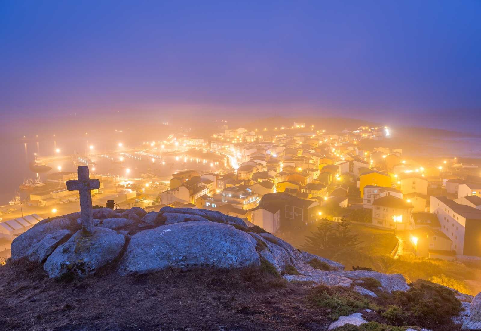 Cruz de piedra sobre una roca dominando un pueblo marinero iluminado entre la niebla al anochecer, etapa de Camariñas a Muxía.